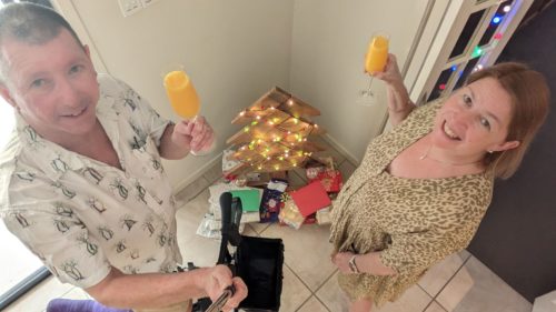 Man and woman with glasses of mimosa in front of small Christmas tree & presents