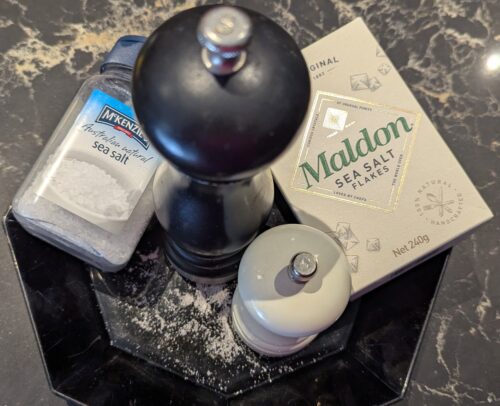 Black plate with a large black and a small white salt cellars, a pot of sea salt and box of Maldon salt flakes, and freshly ground salt on plate.